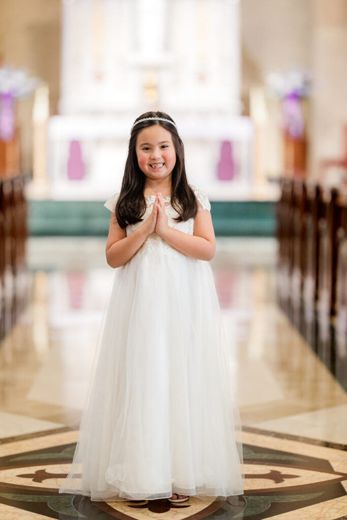 female second grader in white first communion gown posing with prayer hands in front of a traditional catholic altar in roswell, ga