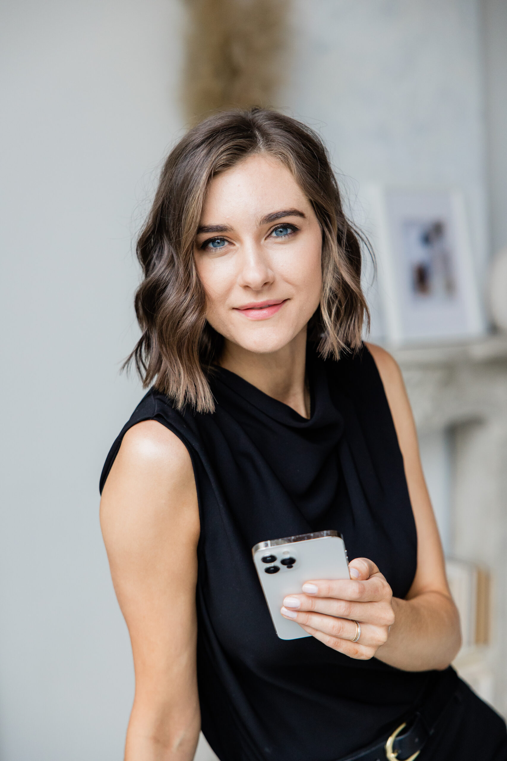 Woman in a black sleeveless top holding a smartphone and smiling, framed in soft studio light.