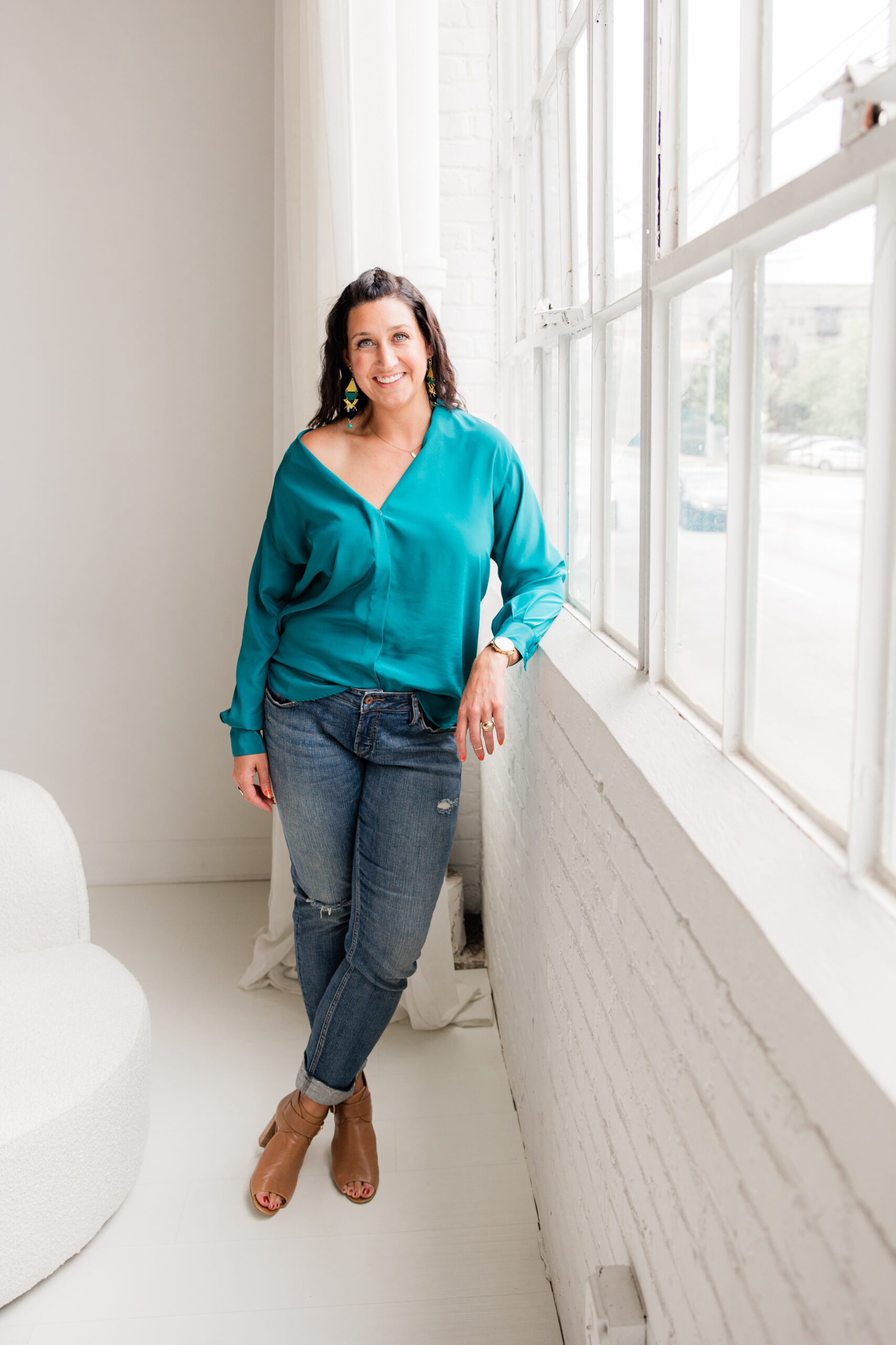 Woman in a teal blouse, jeans, and tan heels leaning against loft windows during an Atlanta branding photoshoot.