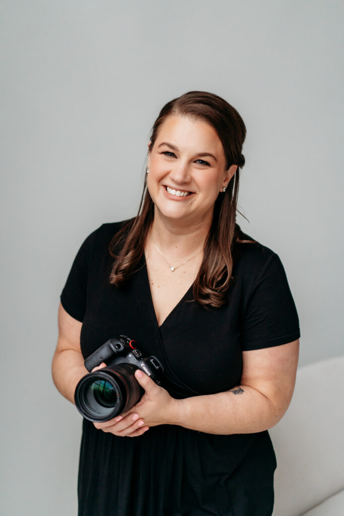 Photographer in a black dress holding her camera and smiling against a simple gray backdrop. Elizabeth Burgler photography can help you decide What to Wear to a Branding Photoshoot