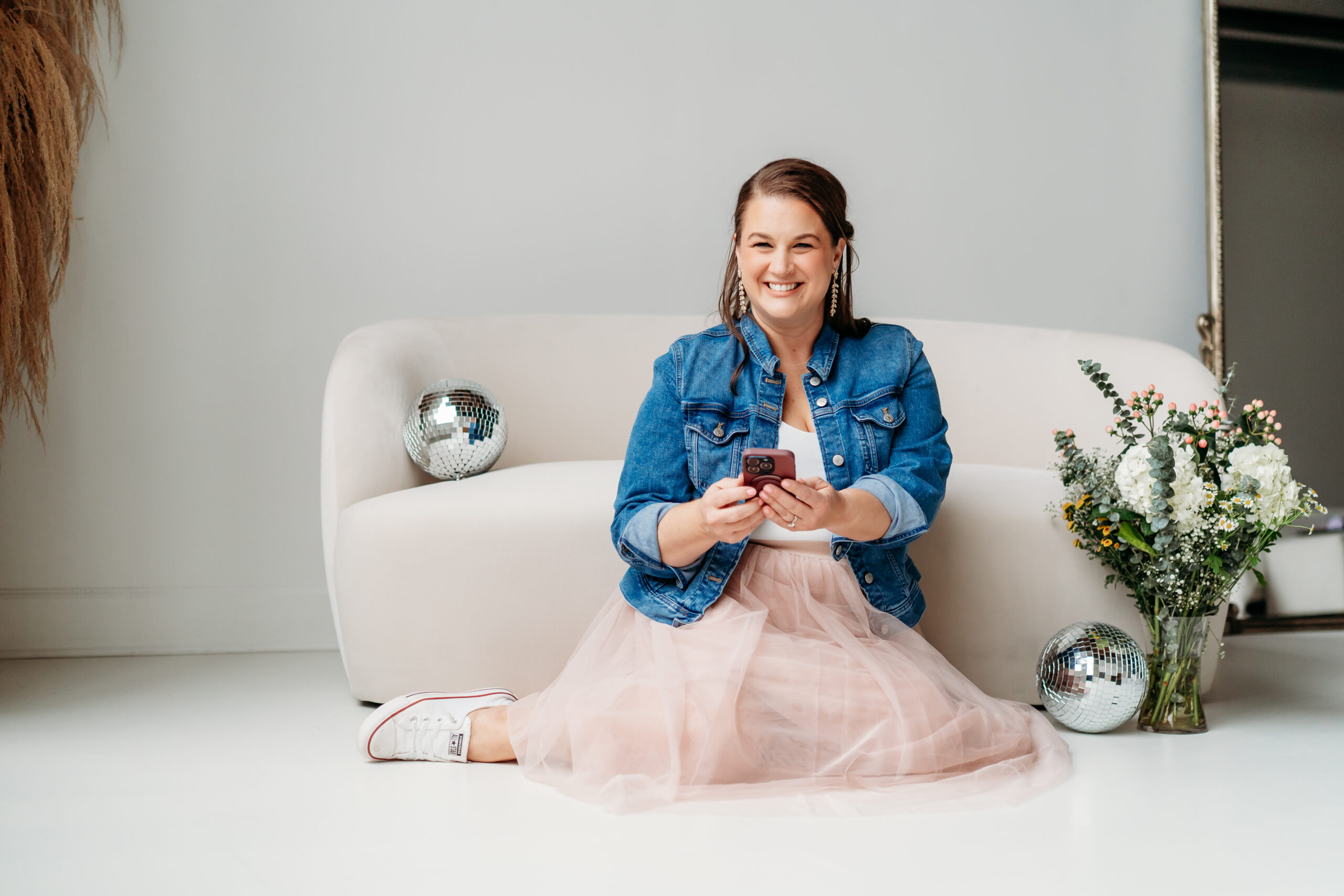 Elizabeth Burgler brand photographer in a tulle skirt, denim jacket, and sneakers sitting on the studio floor with her phone, flowers, and disco balls. Elizabeth help you find What to Wear to a Branding Photoshoot