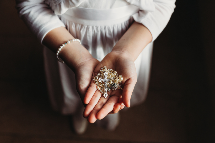 Child's hands holding a rosary during First Communion, captured by a Catholic photographer
